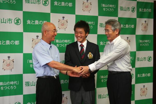 河野君、市長、Yシャツ姿の男性の3人が中央で握手をしている写真